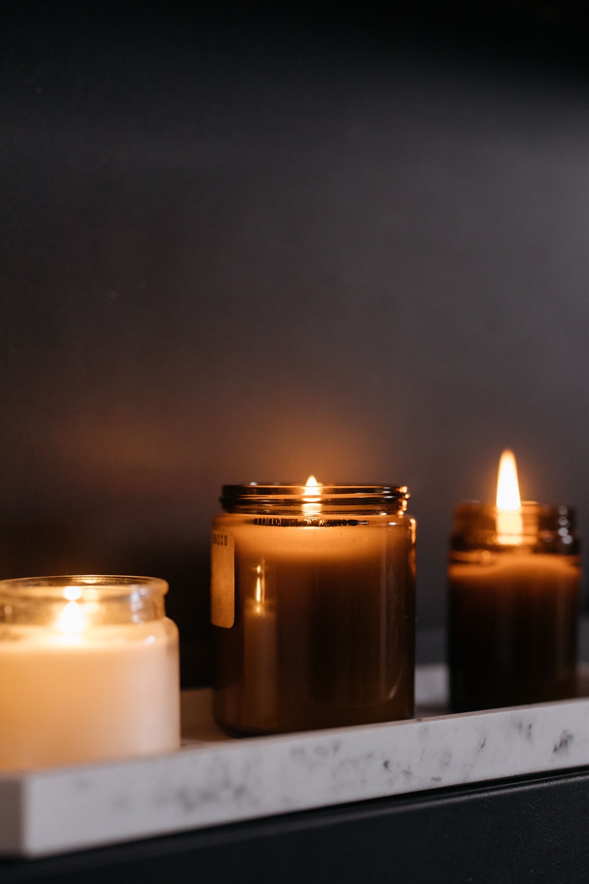 Three lit candles in glass jars arranged on a white marble tray against a dark background. The center candle is a large amber-brown glass jar with a visible flame, flanked by a cream-colored candle on the left and a darker brown candle on the right.