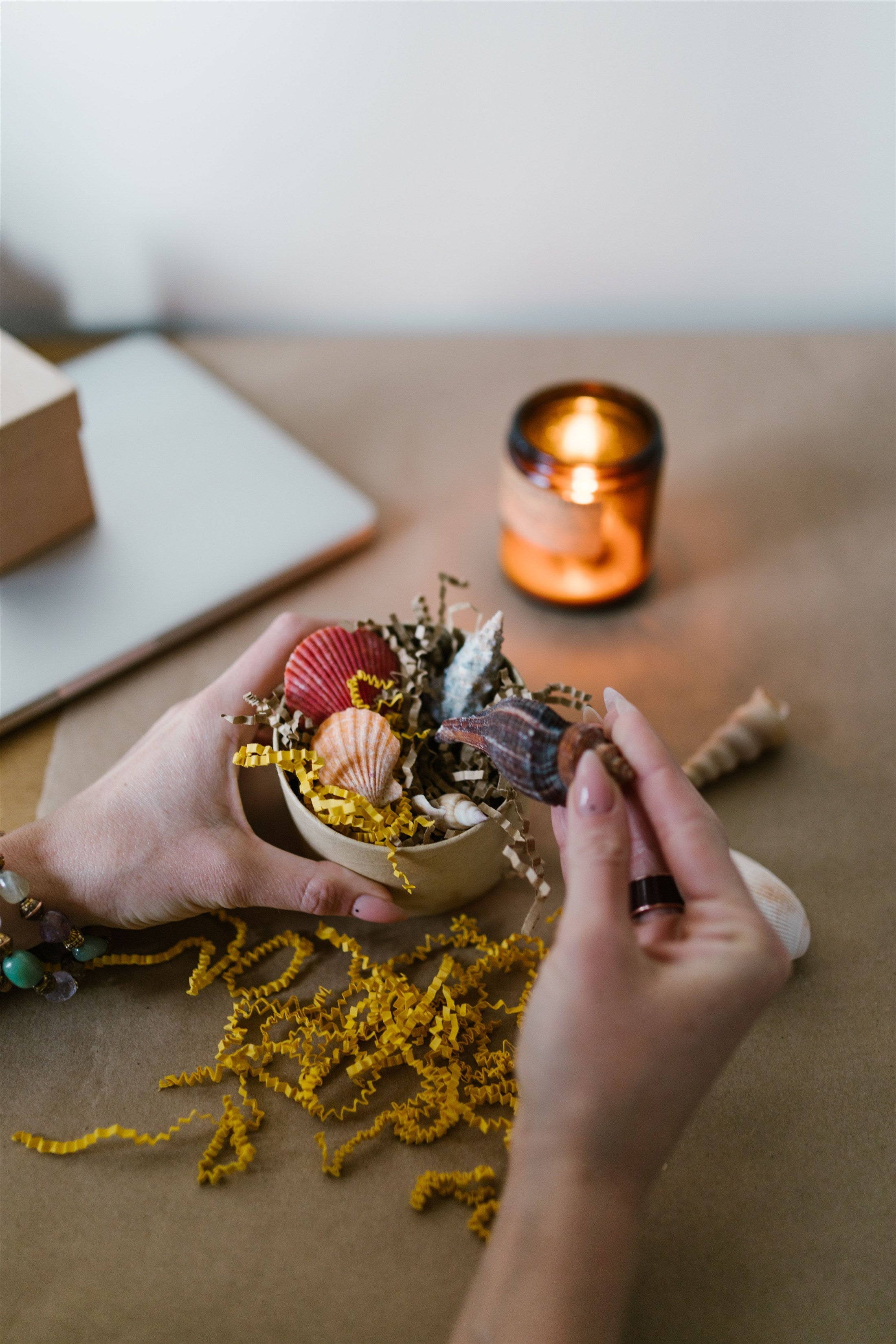 A close-up lifestyle shot of hands arranging a small decorative bowl filled with seashells, dried botanicals, and yellow beads on a kraft paper surface, with a lit candle and laptop blurred in the background.